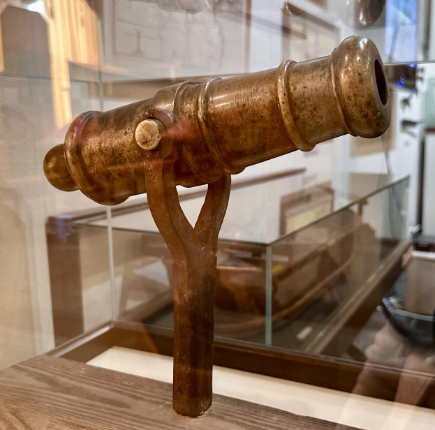 Alte Messingkanone in einer Glasvitrine auf einer hölzernen Unterlage ausgestellt, mit gerahmten Gegenständen an der Wand im Hintergrund.