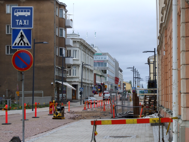 Stadtstraße mit Gebäuden, Straßeninfrastruktur, Fahrzeugen, Bäumen und einer Baustelle mit Verkehrszeichen unter einem bewölkten Himmel.