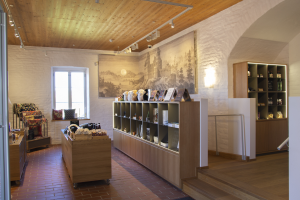 Ein Raum voller verschiedener Möbelstücke, darunter ein Tisch mit Gegenständen, Regalen mit Gegenständen, ein Foto auf der Wand, ein Fenster, Deckenleuchten und eine Treppe, Teil eines neuen Ladens in Heidelberg, Deutschland.