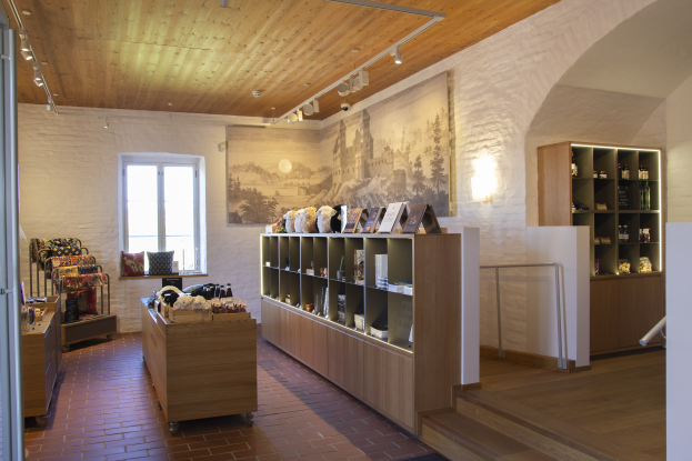 Ein Raum voller verschiedener Möbelstücke, darunter ein Tisch mit Gegenständen, Regalen mit Gegenständen, ein Foto auf der Wand, ein Fenster, Deckenleuchten und eine Treppe, Teil eines neuen Ladens in Heidelberg, Deutschland.
