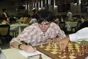 Eine Hand eines Mannes bewegt ein Schachspiel auf einem Brett, während er mit einem Stift schreibt, mit Menschen im Hintergrund.