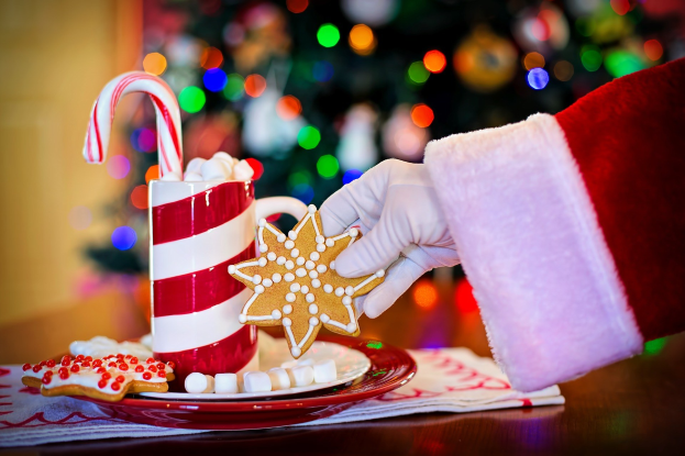 Mann in einem Santa Claus Hut, der eine Tasse heißer Schokolade und einen Zuckerstängel hält und ein Teller mit Plätzchen und ein Tuch auf dem Tisch vor ihm hat, vor einem Weihnachtsbaum und einer Wand.