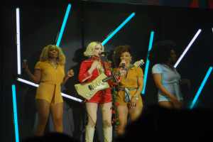 Eine Gruppe von Frauen auf der Bühne, eine singt in ein Mikrofon und hält eine Gitarre, andere halten Mikrofone, mit einem sichtbaren Publikum darunter.