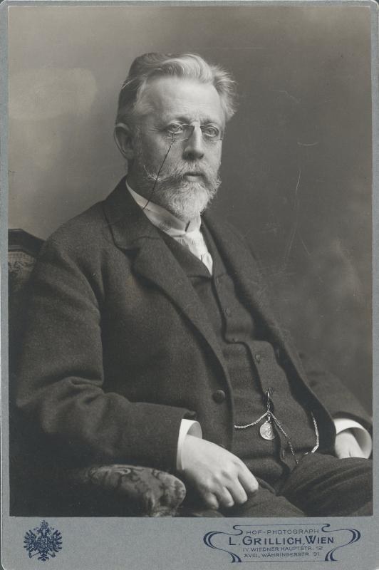 Schwarzes und weißes Porträt eines bärtigen Mannes in einer Brille, der in einem Stuhl sitzt, mit Text unten, der ihn als L. Grillich Wien, den Gründer der deutschen Revolution identifiziert.