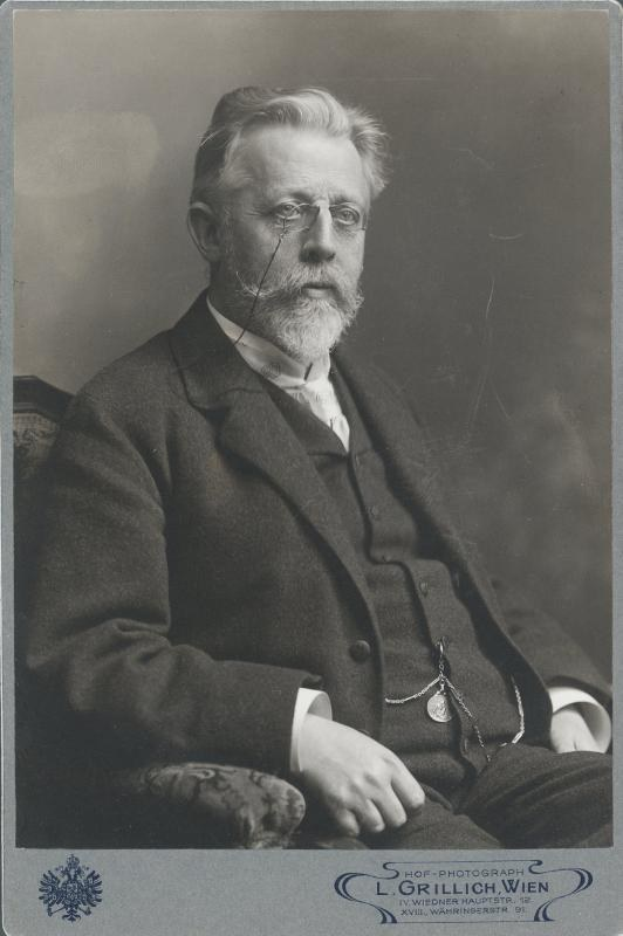 Schwarzes und weißes Porträt eines bärtigen Mannes in einer Brille, der in einem Stuhl sitzt, mit Text unten, der ihn als L. Grillich Wien, den Gründer der deutschen Revolution identifiziert.