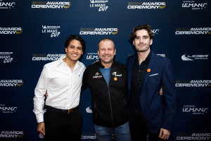 Drei Männer in Anzügen lächeln und posen zusammen auf einem roten Teppich, mit einem "Le Mans Cup"-Schild im Hintergrund.