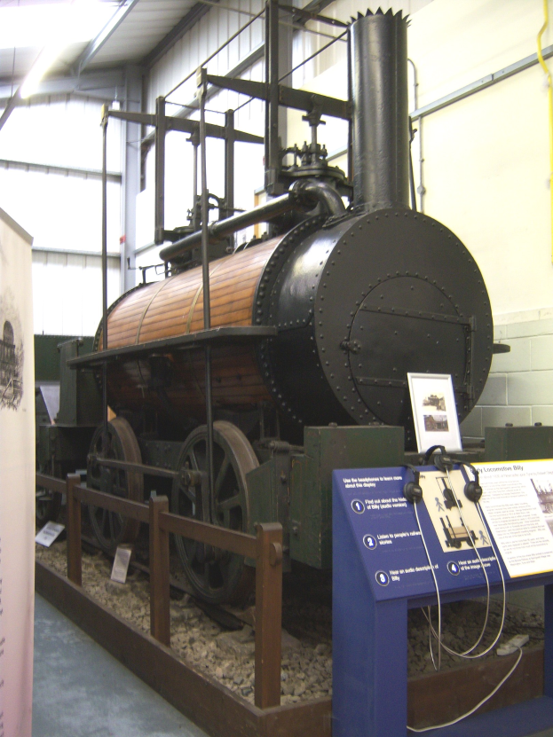 Alte Dampflokomotive in einem Museum ausgestellt, umgeben von Informationsschildern, einem Zaun, Steinen, einer Fahne, einer Wand mit Rohren und Deckenleuchten.