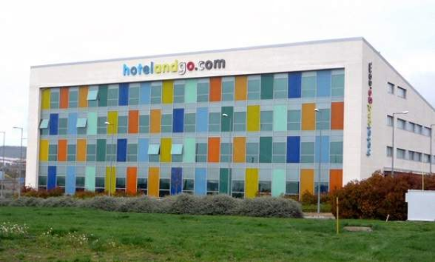 Großes modernes Hotelgebäude mit zahlreichen Fenstern, umgeben von Grünflächen, einem Zaun, Straßenlaternen, geparkten Fahrzeugen, fernen Hügeln und einem bewölkten Himmel; die Wörter "hotel und go" sind auf dem Gebäude zu sehen.