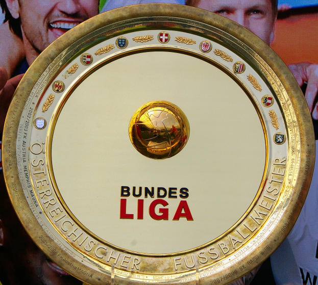 Ein Mann hält einen goldenen Schild mit der Aufschrift "Bundesliga" und eine Fahne mit drei Personen im Hintergrund.