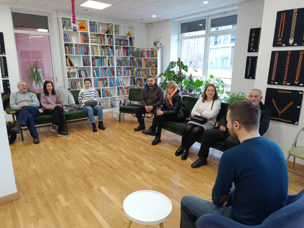 Eine Gruppe von Menschen sitzt um einen Tisch in einem Raum mit Bücherregalen, Pflanzen und Deckenleuchten und ist in ein Gespräch vertieft während eines Workshops zur Zukunft von Gesundheit und Wohlbefinden in Berlin, Deutschland.