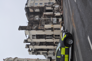 Ein Polizeiwagen steht vor einem zerstörten Gebäude mit zersplitterten Fenstern und verstreutem Schutt, mit anderen Gebäuden und dem Himmel im Hintergrund.