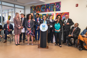 Gruppe von Menschen vor einem Rednerpult mit Mikrofon und einer Tafel mit der Aufschrift "Sancho City Councilman Joins City Council", mit sitzenden und stehenden Teilnehmern, Flaggen, Feuerlöscher und Deckenleuchten.