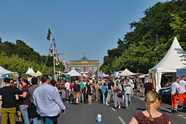 Eine Menschenmenge geht eine Straße mit Zelten, Fahrzeugen und Bäumen entlang auf einen Bogen unter einem klaren blauen Himmel zu, mit Fahnenmästchen links, was auf das Oktoberfest in München, Deutschland, hindeutet.