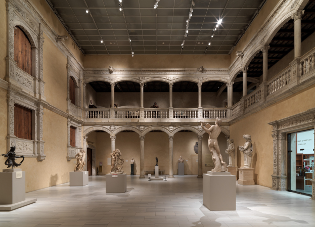 Ein geräumiger Museumsgalerie mit zahlreichen Statuen und Skulpturen auf Sockeln, mit architektonischen Elementen wie Säulen, Geländern und einer beleuchteten Decke und einigen Besuchern, die die Kunstwerke betrachten.