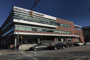 Großes Gebäude mit der Aufschrift "New York City Health Care Homeless Center" mit parkenden Autos davor, umgeben von Straßeninfrastruktur, Fußgängern auf dem Gehweg und Bäumen unter einem klaren blauen Himmel.
