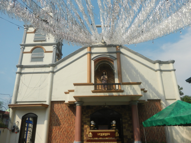Weiße Kirche mit grünem Zelt davor, umgeben von Bäumen unter einem klaren blauen Himmel, mit dekorativen Gegenständen, die von der Kirchturmspitze hängen.