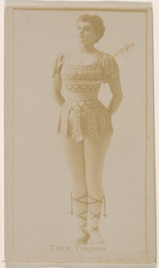 Ein altes Foto einer Frau in einem hellfarbenen gemusterten Badeanzug, ihre Haare zu einem Dutt gebunden, die direkt in die Kamera schaut und lächelt, mit ihren Händen vor der Brust verschränkt.