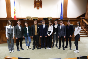 Gruppe von strahlenden Menschen vor einem Gerichtssaal mit Monitoren und Mikrofonen auf Tischen, einer hölzernen Plattform mit Stühlen, Flaggen und einem an der Wand montierten Objekt im Hintergrund und einer Treppe mit Geländern auf der rechten Seite.