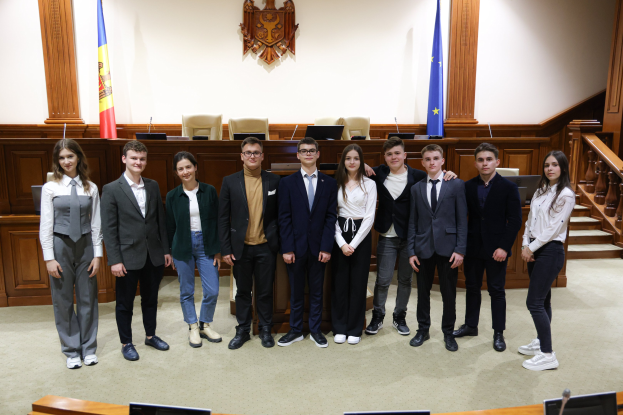 Gruppe von strahlenden Menschen vor einem Gerichtssaal mit Monitoren und Mikrofonen auf Tischen, einer hölzernen Plattform mit Stühlen, Flaggen und einem an der Wand montierten Objekt im Hintergrund und einer Treppe mit Geländern auf der rechten Seite.