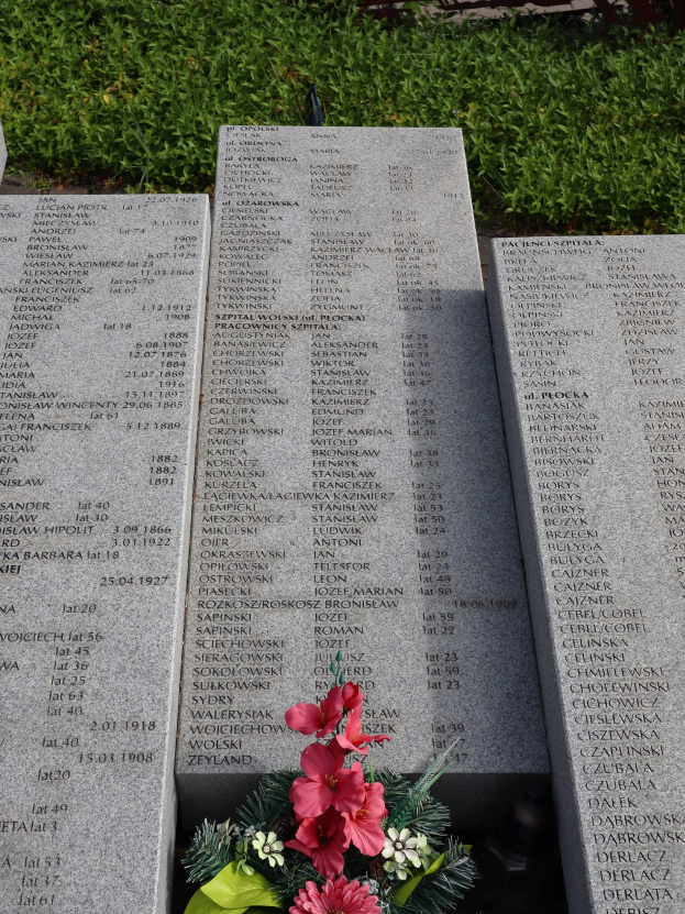 Denkmalstein mit einem Blumenstrauß darauf, umgeben von grünem Gras, mit Text, der wahrscheinlich Kriegsopfer gedenkt.