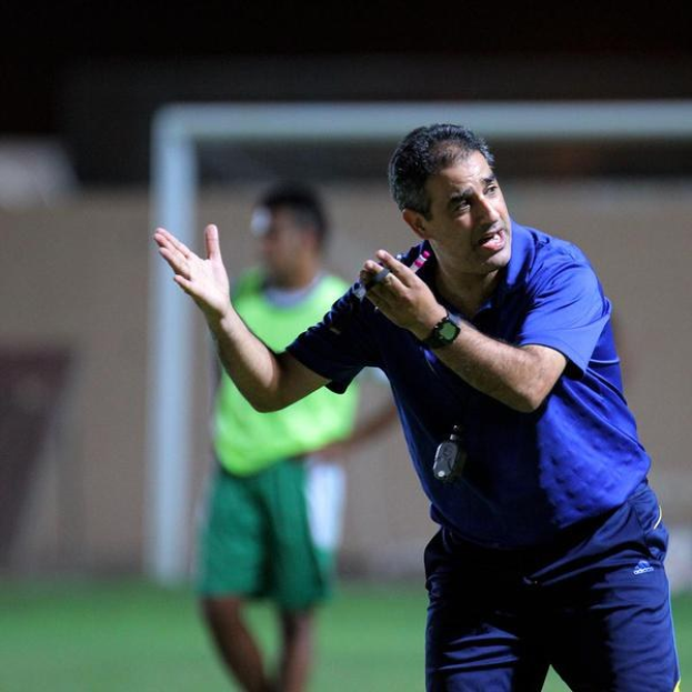 Ein Mann in einem blauen T-Shirt und Hose steht auf einem Fußballfeld und hält ein Mikrofon in der Hand, während er als Trainer gestikuliert. Im Hintergrund ist eine Person in einem grünen Kleid, ein Torpfosten und eine Wand zu sehen.