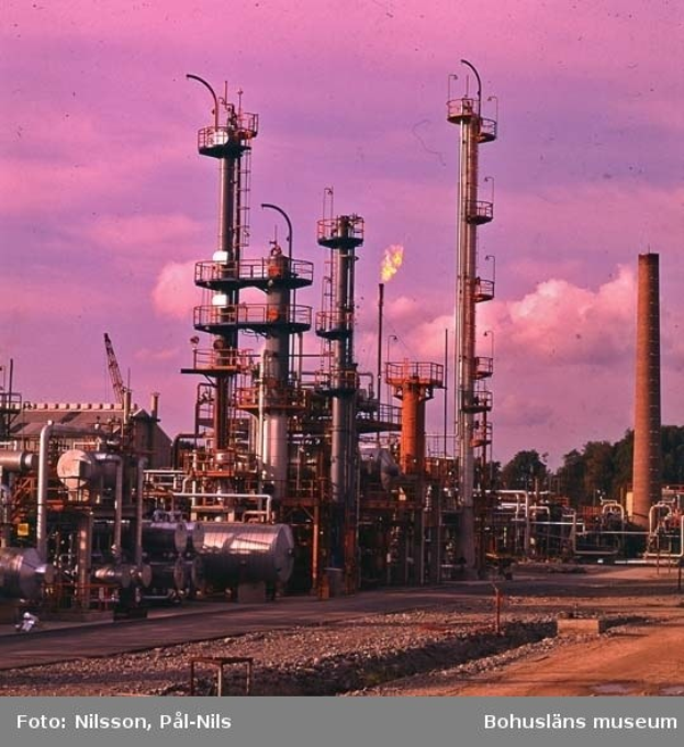 Industrielle Ölfabrik mit ausgedehnten Rohrleitungen, Metallstrukturen, Lagerbehältern, Bäumen, einem Gebäude, einem Kran und einem bewölkten Himmel, mit Text unten.