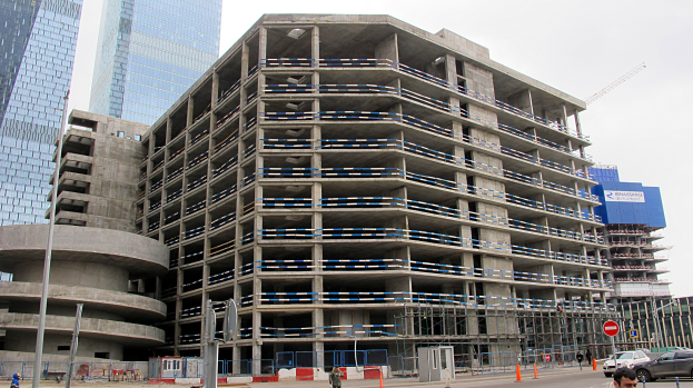 Großes Gebäude im Bau in einer Stadt umgeben von Wolkenkratzern, mit Fahrzeugen, Fußgängern, Verkehrsinfrastruktur und Kränen unter einem klaren blauen Himmel.