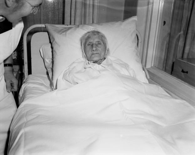 Eine ältere Frau liegt in einem Krankenhausbett mit einer weißen Decke und einem Kissen, während eine Krankenschwester neben ihr steht, mit einem Fenster und Vorhängen im Hintergrund.