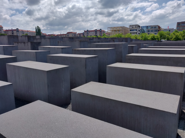 Konkreter Holocaust-Denkmal mit rechteckigen Blöcken, die in einem Gittermuster angeordnet sind, umgeben von Bäumen und Gebäuden unter einem bewölkten Himmel.
