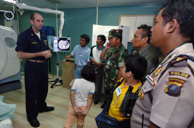 Eine Gruppe von Menschen in einem Krankenhauszimmer mit einem Mann, der ein Mikrofon hält, einem Bett mit einer Decke, medizinischer Ausrüstung, einem Röntgengerät, einer Tafel mit Text, einer Tür und Deckenleuchten.