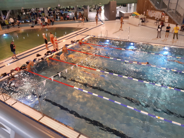 Großes Hallenbad mit schwimmenden Menschen, anderenstehenden, sitzenden oder Gegenstände haltenden Personen rechts von Treppen und Geländern sowie einer Wand im Hintergrund.
