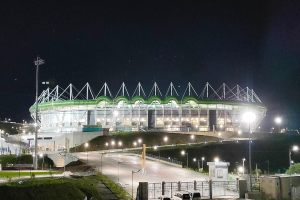 Ein großes Stadion, das nachts beleuchtet ist, mit parkenden Autos davor, umgeben von Straßenmasten, Straßenlaternen, Strommasten, Stromkabeln, Kraftfahrzeugen auf der Straße, Grills, Bäumen und einem Sternenhimmel.