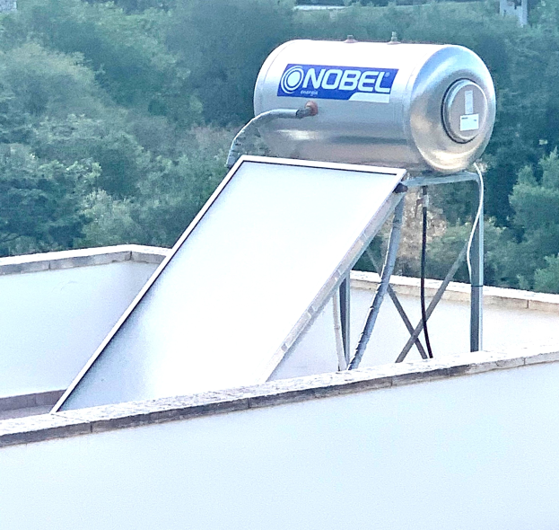 Solarwassererhitzer mit Speicherbehälter auf dem Dach eines Gebäudes montiert, Bäume im Hintergrund sichtbar.