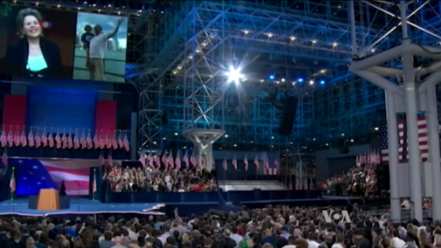 Eine große Menschenmenge sitzt vor einer Bühne mit einem Podium, einem Mikrofon, Flaggen, einem Bildschirm und Beleuchtungsausrüstung und wartet auf eine Präsidentschaftsdebatte.