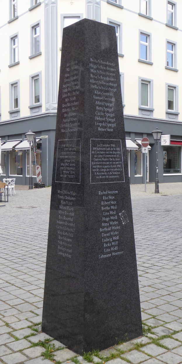 Ein Denkmal mit Inschrift steht auf einem Stadtplatz umgeben von Gebäuden, Straßenmöbeln und Menschen unter einem sichtbaren Himmel.