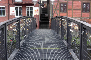 Eine Brücke, geschmückt mit Liebesschlössern, spannt sich vor einem Backsteingebäude, beleuchtet von Laternen, mit Stufen, die zum Gebäude hinaufführen, und Text am unteren Bildrand.