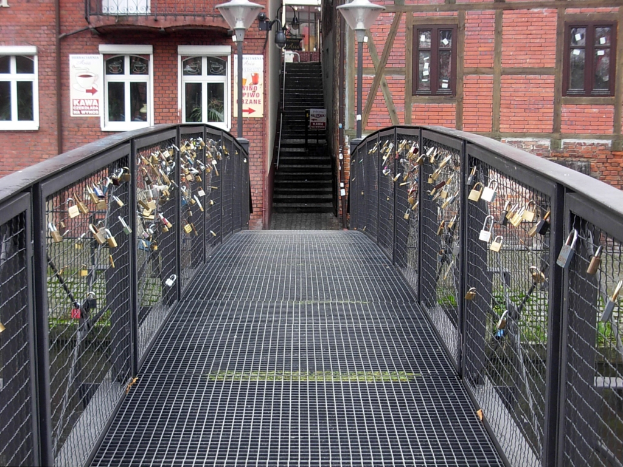 Eine Brücke, geschmückt mit Liebesschlössern, spannt sich vor einem Backsteingebäude, beleuchtet von Laternen, mit Stufen, die zum Gebäude hinaufführen, und Text am unteren Bildrand.