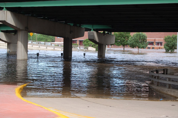 Überschwemmte Stadtstraße mit Wasser bis zur Brückenhöhe, die Straße unter Wasser, mit einem Geländer auf der rechten Seite und Bäumen und Gebäuden im Hintergrund.