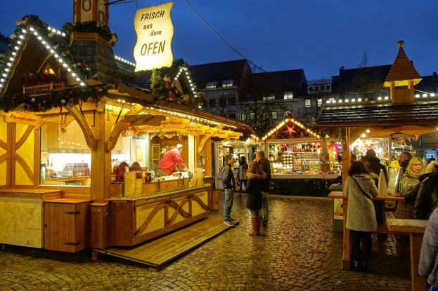 Ein lebendiger Weihnachtsmarkt auf Kopfsteinpflasterstraße bei Nacht, mit Menschen, die an Ständen mit Lichtern und festlicher Dekoration vorbeischlendern, Gebäuden, Bäumen und einem klaren Nachthimmel im Hintergrund.