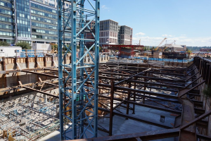 Das Bild zeigt eine Baustelle mit Gebäuden und Büros links, Eisenstangen und Maschinen rechts und Kränen und Geräten in der Mitte, unter einem Himmel in der oberen rechten Ecke.