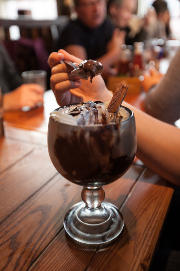 Eine Gruppe von Menschen sitzt um einen Tisch mit einem Glas Schokoladenpudding in der Mitte, eine Person im Vordergrund isst mit einem Löffel, Hintergrund leicht unscharf.