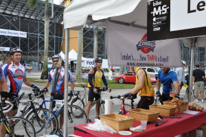 Eine Straßenszene mit Menschen, die in der Nähe von Fahrrädern stehen, ein Geschäft mit Gegenständen auf einem Tisch, Bäume, Zelte, an einem Schuppen befestigte Banner, Fahrzeuge und Gras.