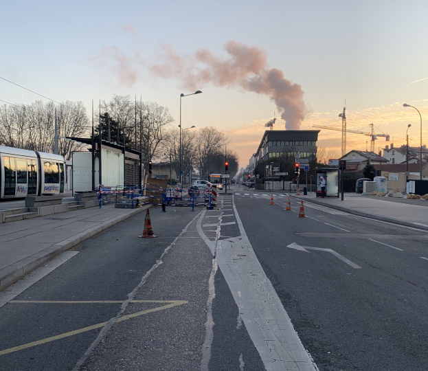 Eine Stadtstraße mit Verkehrskegeln, einem Bus, Laternenmasten, Bäumen, Gebäuden, Kränen und Rauch, der aus einem entfernten Gebäude aufsteigt.