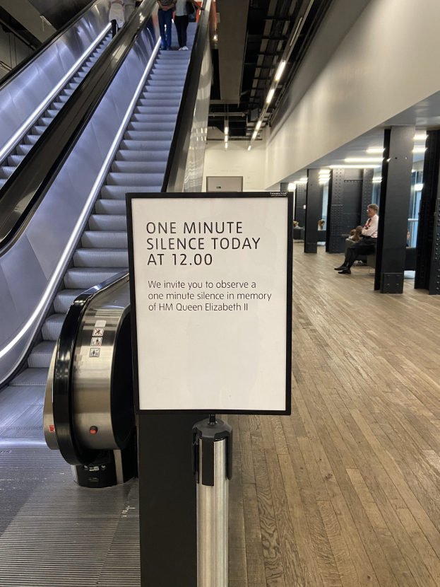 Eine Rolltreppe im Flughafen mit einem Schild, auf dem "Eine Minute Stille heute" steht, sowie ein paar Menschen darauf und an der Decke befestigte Lampen im Hintergrund.
