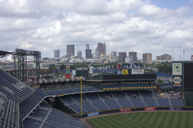 Ein Baseballstadion mit leeren Sitzplätzen, Pfosten und Beschriftungen, vor einer Stadtkulisse unter einem bewölkten Himmel, mit Bäumen und fernen Gebäuden im Hintergrund.