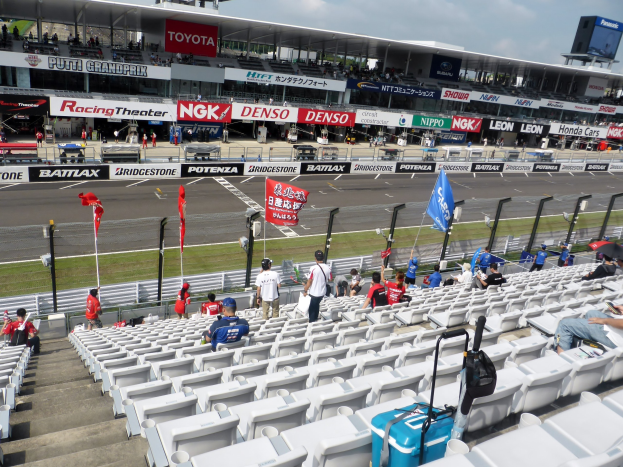 Eine Gruppe von Menschen auf Tribünen bei einer Rennstrecke mit Fahnen, Texttafeln, Geländern und Stufen, mit einem bewölkten Himmel über dem Stadion im Hintergrund.