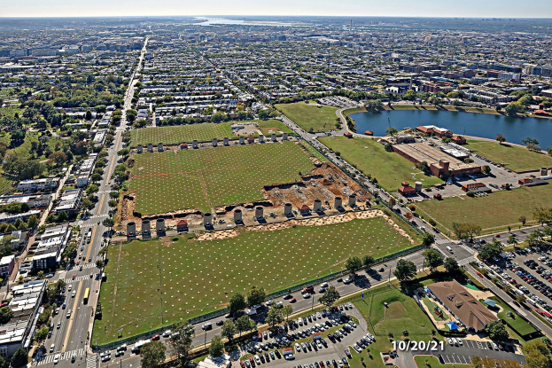 Luftaufnahme einer Stadt mit dichtem Baumbestand, Grünflächen, Straßen mit Fahrzeugen, einem Gewässer und einem bewölkten Himmel, die einen großen zentralen Platz für einen geplanten Park zeigt.