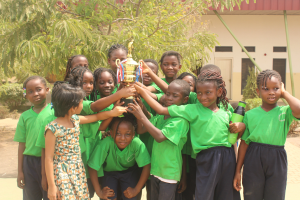 Eine Gruppe von Kindern, die gemeinsam mit einem Pokal posen, umgeben von Bäumen, Pflanzen und einem Gebäude.