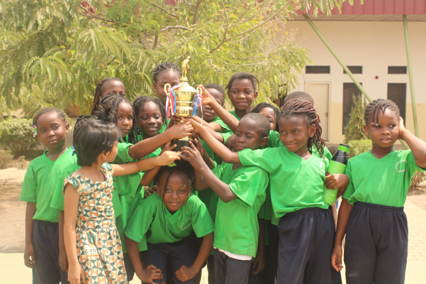 Eine Gruppe von Kindern, die gemeinsam mit einem Pokal posen, umgeben von Bäumen, Pflanzen und einem Gebäude.
