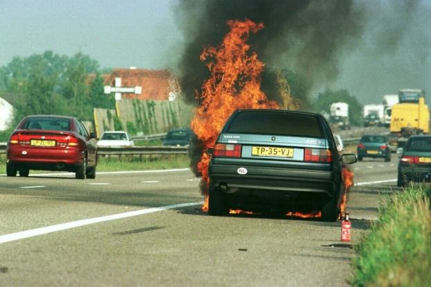 Ein Auto in Flammen an der Seite der Straße, umgeben von anderen Fahrzeugen, mit Bäumen, Gebäuden und einem klaren blauen Himmel im Hintergrund und einem Feuerlöscher auf der rechten Seite.